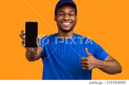 Recommending Delivery Service. Smiling african american male courier holding smartphone with blank empty screen for mockup and showing thumbs up gesture, wearing blue cap and uniform, orange studio Recommending Delivery Service. Smiling african american male courier holding smartphone with blank empty screen for mockup and showing thumbs up gesture, wearing blue cap and uniform, orange studio 135503695