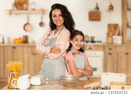 Cooking Team. Smiling Arabic Mom And Her Daughter Enjoying Baking Leisure In Modern Kitchen Indoor, Standing Back To Back Near Table, Posing Ready To Make Tasty Pastry, Cake and Cookies Together 135503722