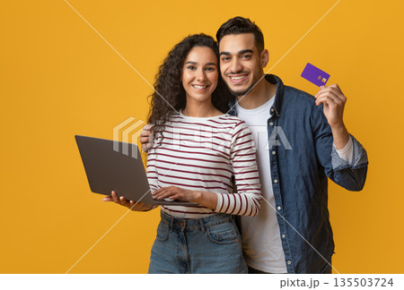 Online Shopping Concept. Happy Arabic Couple Using Credit Card And Laptop While Standing Over Yellow Background In Studio, Cheerful Middle Eastern Man And Woman Enjoying Purchasing In Internet Online Shopping Concept. Happy Arabic Couple Using Credit Card And Laptop While Standing Over Yellow Background In Studio, Cheerful Middle Eastern Man And Woman Enjoying Purchasing In Internet 135503724