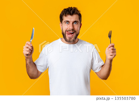 Hungry young guy holding knife and fork looking at camera, posing on yellow studio background. Portrait of funny young man having food cravings and temptation ready for tasty dinner 135503780