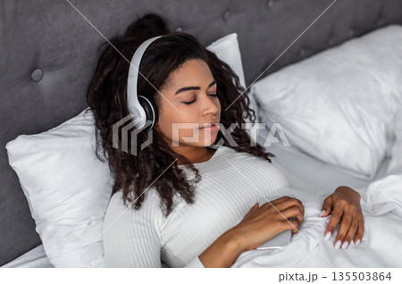 Rest And Relax Concept. Closeup portrait of calm black woman lying in bed, listening to music, audio book, podcast, enjoying meditation for sleep and peaceful mind in wireless headphones. Zen-like Rest And Relax Concept. Closeup portrait of calm black woman lying in bed, listening to music, audio book, podcast, enjoying meditation for sleep and peaceful mind in wireless headphones. Zen-like 135503864