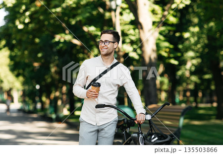 Healthy and active lifestyle, going to work on eco transport. Smiling attractive millennial male employee in glasses with bag and cup of takeaway coffee, moving bike in city park in summer, outdoor 135503866