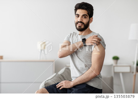 Recommendations for vaccination and immunization of population, health care concept. Cheerful arabian bearded young male shows hand with band aid after injection in living room interior, empty space Recommendations for vaccination and immunization of population, health care concept. Cheerful arabian bearded young male shows hand with band aid after injection in living room interior, empty space 135503879