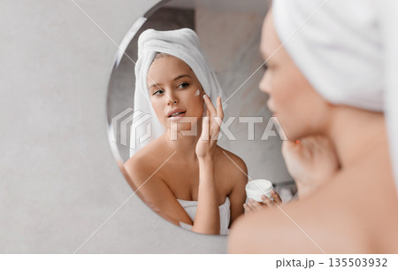 Home beauty routine. Young woman applying moisturising cream on face looking at miror in bathroom, free space. Attractive lady wearing white towel on head after shower 135503932