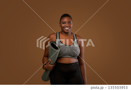 Ready For Training. Portrait of African American chubby woman in bra top and leggings going to gym, holding carrying roll mat standing isolated over brown studio background, looking posing at camera Ready For Training. Portrait of African American chubby woman in bra top and leggings going to gym, holding carrying roll mat standing isolated over brown studio background, looking posing at camera 135503953