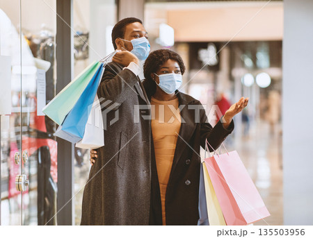 Seasonal sales during coronavirus pandemic. Happy african american family in coats and protective masks walking in mall after buying clothes at Black Friday, lady showing at store window, copy space Seasonal sales during coronavirus pandemic. Happy african american family in coats and protective masks walking in mall after buying clothes at Black Friday, lady showing at store window, copy space 135503956