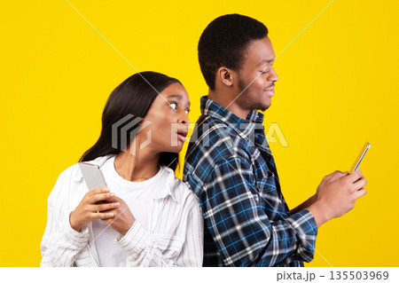 Jealousy, online networks and relationships. Interested millennial african american student lady in casual looks into phone of smiling husband, isolated on yellow background, empty space, studio shot Jealousy, online networks and relationships. Interested millennial african american student lady in casual looks into phone of smiling husband, isolated on yellow background, empty space, studio shot 135503969