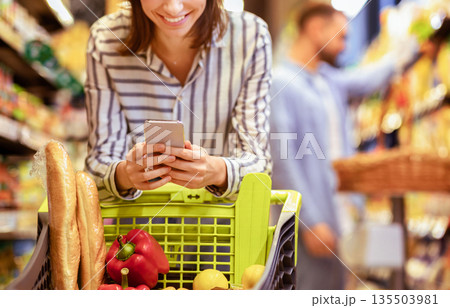 Couple In The Supermarket. Cropped closeup image of unrecognizable smiling woman leaning on shopping cart, using mobile phone, her boyfriend is choosing food in the blurred background 135503981