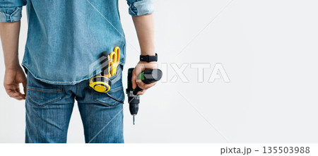 Men work, home renovation, construction or carpentry business. Young guy in jeans holds electric drill, with tools in his pocket, isolated on white background, panorama, empty space, cropped, back 135503988