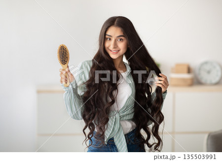 Portrait of happy Indian lady brushing her long dark hair with wooden brush at home. Lovely young woman taking care of her beautiful locks. Beauty, wellness and hairdressing concept 135503991