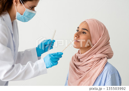 PCR Test. Nurse Taking Swab From Muslim Female Patient's Nose, Lady Doctor In Medical Mask Making Covid-19 Diagnostic Procedure To Islamic Woman In Hijab Over White Background, Closeup Shot 135503992