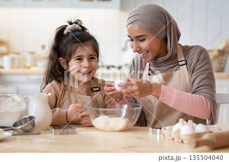 Happy islamic mom and her cute daughter preparing dough in kitchen together, cracking eggs to bowl, enjoying cooking homemade food, little girl and her muslim mother in hijab having fun at home Happy islamic mom and her cute daughter preparing dough in kitchen together, cracking eggs to bowl, enjoying cooking homemade food, little girl and her muslim mother in hijab having fun at home 135504040