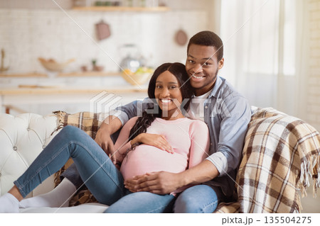 Beautiful pregnant couple hugging on sofa at home. Young African American man embracing his happy expectant wife indoors. Smiling future mom and dad waiting for their baby Beautiful pregnant couple hugging on sofa at home. Young African American man embracing his happy expectant wife indoors. Smiling future mom and dad waiting for their baby 135504275