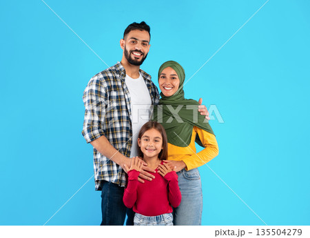 Modern Muslim Family Of Three Hugging Smiling To Camera Standing Together Over Blue Studio Background. Islamic Mother In Hijab And Father Embracing Their Happy Little Daughter Modern Muslim Family Of Three Hugging Smiling To Camera Standing Together Over Blue Studio Background. Islamic Mother In Hijab And Father Embracing Their Happy Little Daughter 135504279