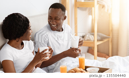 Joyful black young couple sitting in bed in morning and having breakfast, drinking coffee and talking, side view, copy space. Cheerful african american man and woman enjoying breakfast in bed 135504440