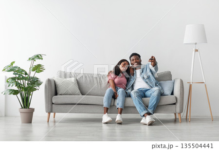 Weekend Leisure. Happy relaxed millennial black guy and lady watching tv in living room interior, holding remote control and hugging. Smiling couple enjoying film and movie evening together Weekend Leisure. Happy relaxed millennial black guy and lady watching tv in living room interior, holding remote control and hugging. Smiling couple enjoying film and movie evening together 135504441
