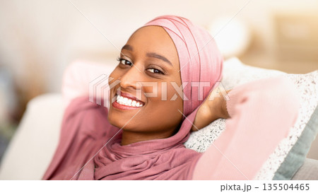 Peaceful weekend morning at home. Happy black lady in hijab relaxing on comfortable sofa indoors. Pretty African American woman in traditional headscarf resting on sofa in living room Peaceful weekend morning at home. Happy black lady in hijab relaxing on comfortable sofa indoors. Pretty African American woman in traditional headscarf resting on sofa in living room 135504465