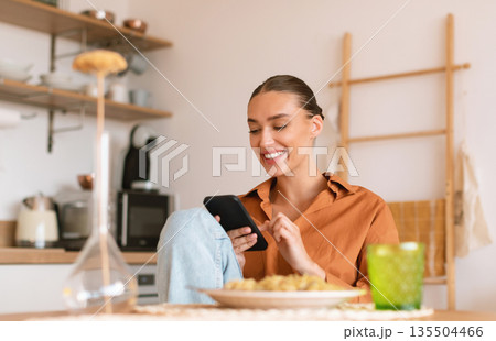 Excited lady using smartphone in kitchen while having dinner, sitting at table and texting on phone, browsing social networks or shopping online, free space Excited lady using smartphone in kitchen while having dinner, sitting at table and texting on phone, browsing social networks or shopping online, free space 135504466