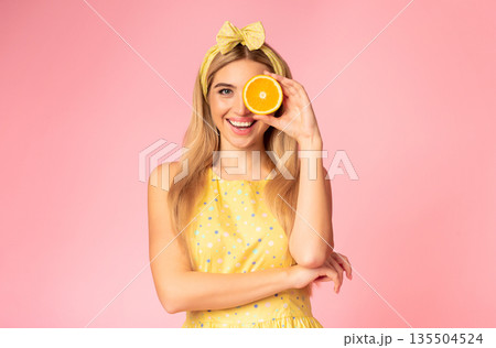 Having Fun. Charming young girl covering eye with orange half, holding piece of fruit near face, copyspace, pink wall Having Fun. Charming young girl covering eye with orange half, holding piece of fruit near face, copyspace, pink wall 135504524