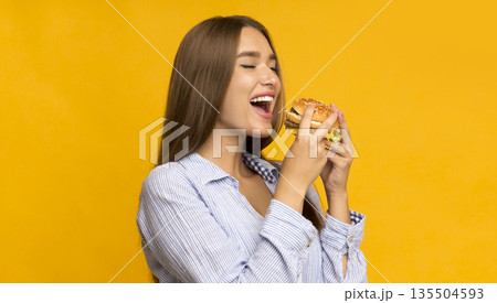 Cheat meal day. Hungry young woman eating burger standing in studio on yellow background. Panorama Cheat meal day. Hungry young woman eating burger standing in studio on yellow background. Panorama 135504593
