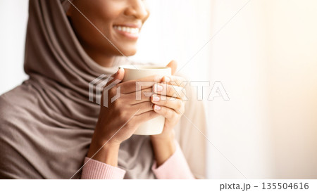 Cropped view of happy black woman in hijab warming her hands on cup of hot coffee near window at home. Lovely young Muslim lady having relaxing morning with aromatic drink, enjoying her break 135504616