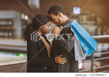 Romance for two, tenderness and date during seasonal sales and Black Friday. Young african american couple in love with colored shopping bags hugging at city mall ready to buy, side view, free space 135504617