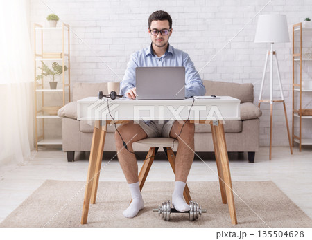 Video call and working from home. Young man in glasses, shirt, shorts and socks at workplace, with dumbbell in interior of living room 135504628