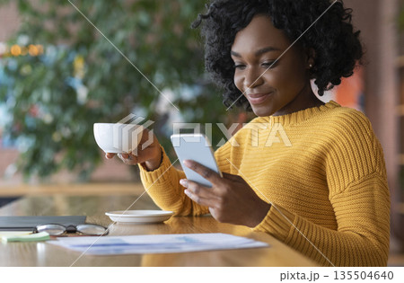 Young afro girl having morning coffee at cafe alone, using phone, close up, empty space Young afro girl having morning coffee at cafe alone, using phone, close up, empty space 135504640