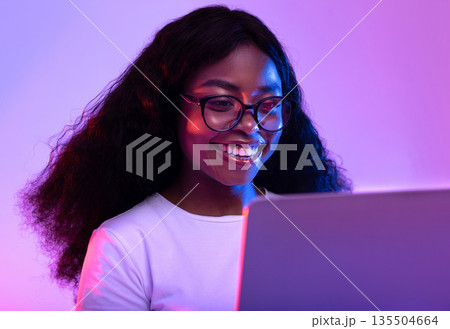 Happy young black woman in glasses using laptop pc for online work or studies in neon light. Cheery African American lady having remote video conference, chatting with her friends on web Happy young black woman in glasses using laptop pc for online work or studies in neon light. Cheery African American lady having remote video conference, chatting with her friends on web 135504664