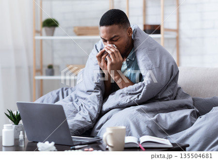 Ill African Man Having Rhinitis Sneezing And Blowing Runny Nose In Paper Tissue Sitting On Sofa At Home, Working Distantly On Laptop. Sinusitis Illness, Cold And Influenza Symptoms. 135504681