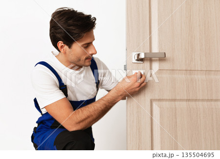 Repair Service Concept. Portrait of smiling handsome young locksmith in blue uniform installing door knob on front wooden entrance door at home or office workplace, isolated on white studio background 135504695