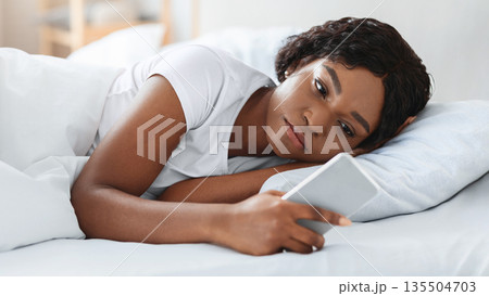 Frustrated young black woman looking at smartphone screen while laying in bed before sleeping, feeling lonely or depressed, closeup. Upset african american lady waiting for call from lover Frustrated young black woman looking at smartphone screen while laying in bed before sleeping, feeling lonely or depressed, closeup. Upset african american lady waiting for call from lover 135504703