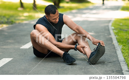 Sports injury. African American runner sitting on jogging track and feeling pain in his ankle, panorama 135504798
