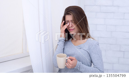 Sad young woman with cup of coffee crying near window, wiping her tears at home. Depressed millennial lady feeling lonely, stressed or desperate, suffering from mental disorder 135504799