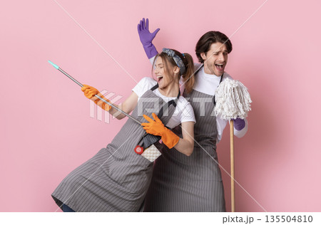 Playful couple of cleaners fooling together while making spring tidying, singing into mop and using broom as guitar over pink background Playful couple of cleaners fooling together while making spring tidying, singing into mop and using broom as guitar over pink background 135504810