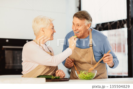 Cheerful Senior Couple Feeding Each Other And Laughing Enjoying Cooking Together In Modern Kitchen At Home. Older Spouses Having Fun Tasting Food. Family Dinner Preparation Cheerful Senior Couple Feeding Each Other And Laughing Enjoying Cooking Together In Modern Kitchen At Home. Older Spouses Having Fun Tasting Food. Family Dinner Preparation 135504829