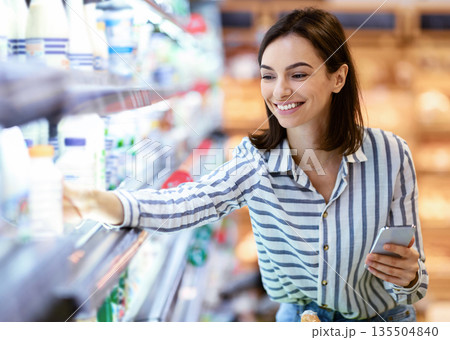 Shopping. Young Smiling Woman Holding And Using Phone Buying Food Groceries Standing In Supermarket. Female Customer With Smartphone Taking Milk From The Fridge In Dairy Section At The Shop 135504840