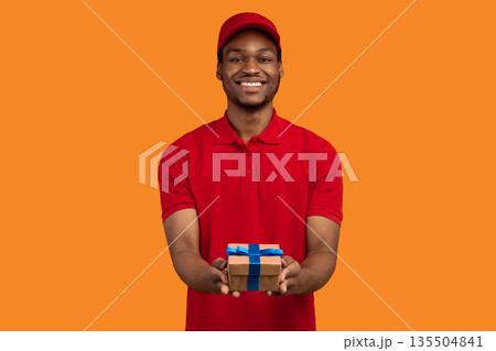 Delivery Service For Holidays. Portrait of smiling cheerful african american courier guy holding wrapped gift box, giving and showing present to camera, isolated over orange studio background 135504841