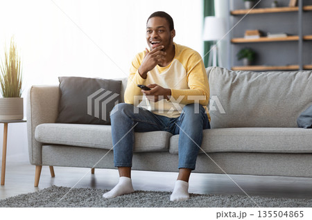 Watching TV at home. Happy african american man having rest, switching channels with remote controller and smiling, empty space. Black guy enjoying his weekend, watching TV in living room 135504865