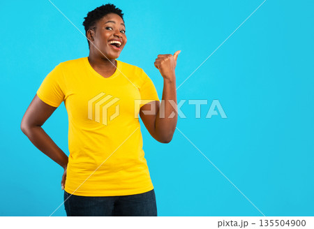 Joyful Plus Size African American Lady Pointing Thumb Finger Aside At Empty Space For Text Recommending Something Standing In Studio Over Blue Background. Look There Advertisement Banner Joyful Plus Size African American Lady Pointing Thumb Finger Aside At Empty Space For Text Recommending Something Standing In Studio Over Blue Background. Look There Advertisement Banner 135504900