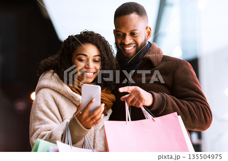 Shopping Together Concept. Portrait of happy african american couple using smartphone, pointing at gadget, holding shopping paper bags, standing outdoors in the evening. Retail, Sale, Technology 135504975