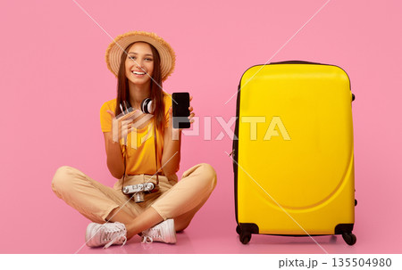 Carefree girl traveller pointing at smartphone blank screen over pink studio background, sitting next to luggage Carefree girl traveller pointing at smartphone blank screen over pink studio background, sitting next to luggage 135504980