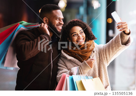 Couple Selfie. Portrait of happy casual african american woman and man taking picture on the street near mall, smiling at camera, holding colorful shopping bags with purchases. Family Photo 135505136