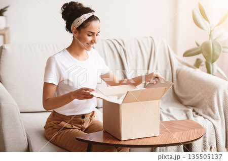 Happy young woman unpacking delivery box, sitting on couch by tea table, home interior. Satisfied customer cheerful lady opening package, ordering clothes or cosmetics on Internet, copy space Happy young woman unpacking delivery box, sitting on couch by tea table, home interior. Satisfied customer cheerful lady opening package, ordering clothes or cosmetics on Internet, copy space 135505137