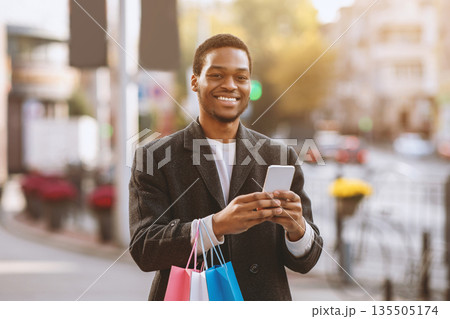 Low price and Black Friday, Seasonal Sale and mobile app. Smiling african american man in coat holding multicolor shopping bags with purchases and smartphone, and rejoices to cashback, free space Low price and Black Friday, Seasonal Sale and mobile app. Smiling african american man in coat holding multicolor shopping bags with purchases and smartphone, and rejoices to cashback, free space 135505174