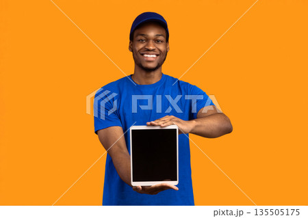 Delivery Application. African american courier man holding digital tablet with blank screen for mock up and showing it to camera, wearing blue cap and uniform, standing isolated on orange background Delivery Application. African american courier man holding digital tablet with blank screen for mock up and showing it to camera, wearing blue cap and uniform, standing isolated on orange background 135505175