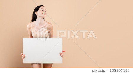 Smiling young female with vitiligo skin disorder holding white blank placard, beautiful millennial lady standing in underwear over beige background in studio, enjoying her different beauty, copy space 135505193