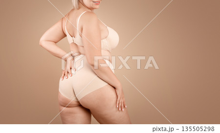 Young plus size woman in beige underwear posing over brown studio background, half-turned at camera, cropped image with free space Young plus size woman in beige underwear posing over brown studio background, half-turned at camera, cropped image with free space 135505298
