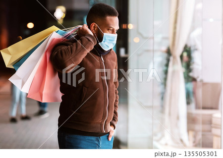 Safe Shopping, Quarantine, Lockdown, Pandemic. Black guy wearing disposable face mask looking at shop window of the shopping center, walking in the evening near mall, carrying colorful bags 135505301