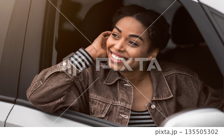 Pleasant looks african american female with glad expression, being satisfied with unforgettable journey by car, sits on driver s seat, enjoys music and look at open window. People, driving, transport 135505302